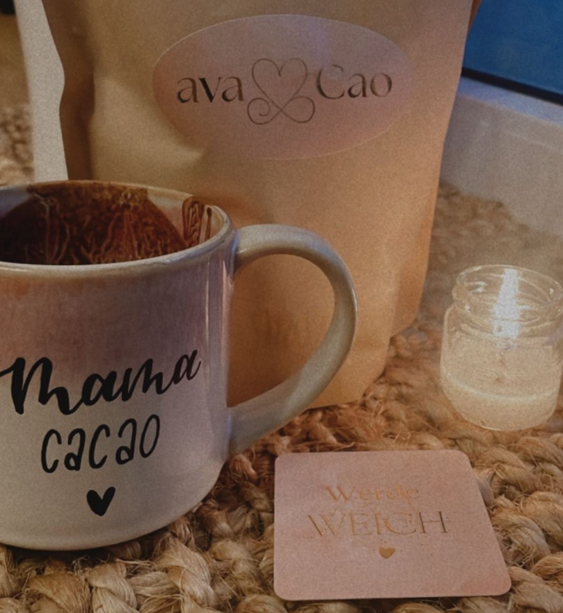 Kakaotasse mit der Aufschrift „Mama Cacao“ steht neben einer avaCao-Verpackung, einer Kerze und einer Karte mit der Botschaft „Werde weich“ auf einem geflochtenen Untersetzer.