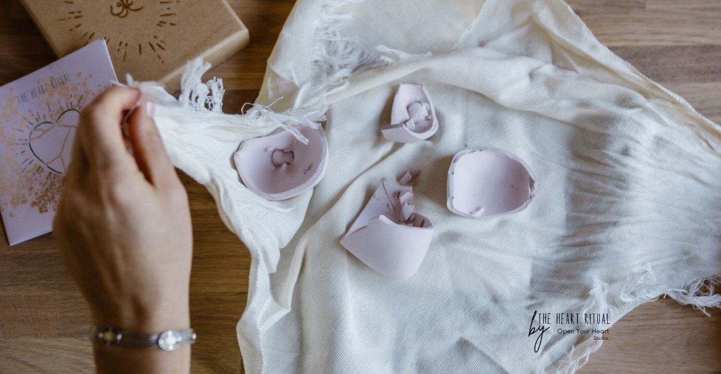 Zerbrochene Keramikteile eines rosa Herzens liegen auf einem Tuch, daneben eine Hand und eine Karte mit Herzsymbol