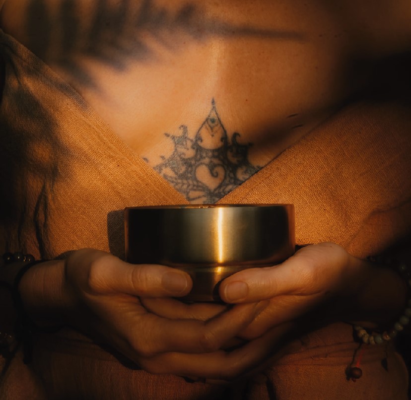 Zwei Hände halten eine goldene Cacaoschale vor einem tätowierten Brustbereich mit spirituellem Symbol.