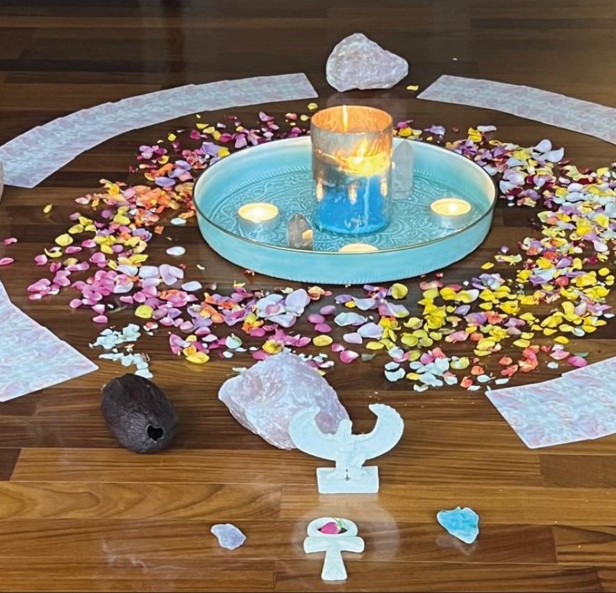 Ein liebevoll gestalteter Zeremonienkreis mit einer brennenden Kerze, Blütenblättern, Cacao-Frucht, Rosenquarzen und spirituellen Symbolen auf einem Holzboden – eine Einladung zur heiligen Verbindung.