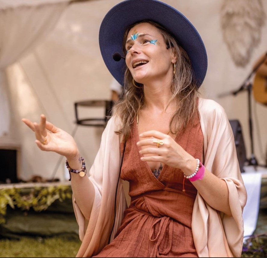 Ava mit Mikrofon, bemaltem Gesicht und weitem Hut in einem farbenfrohen, spirituellen Setting – sie spricht und gestikuliert lebendig inmitten einer Zeremonie oder Session.
