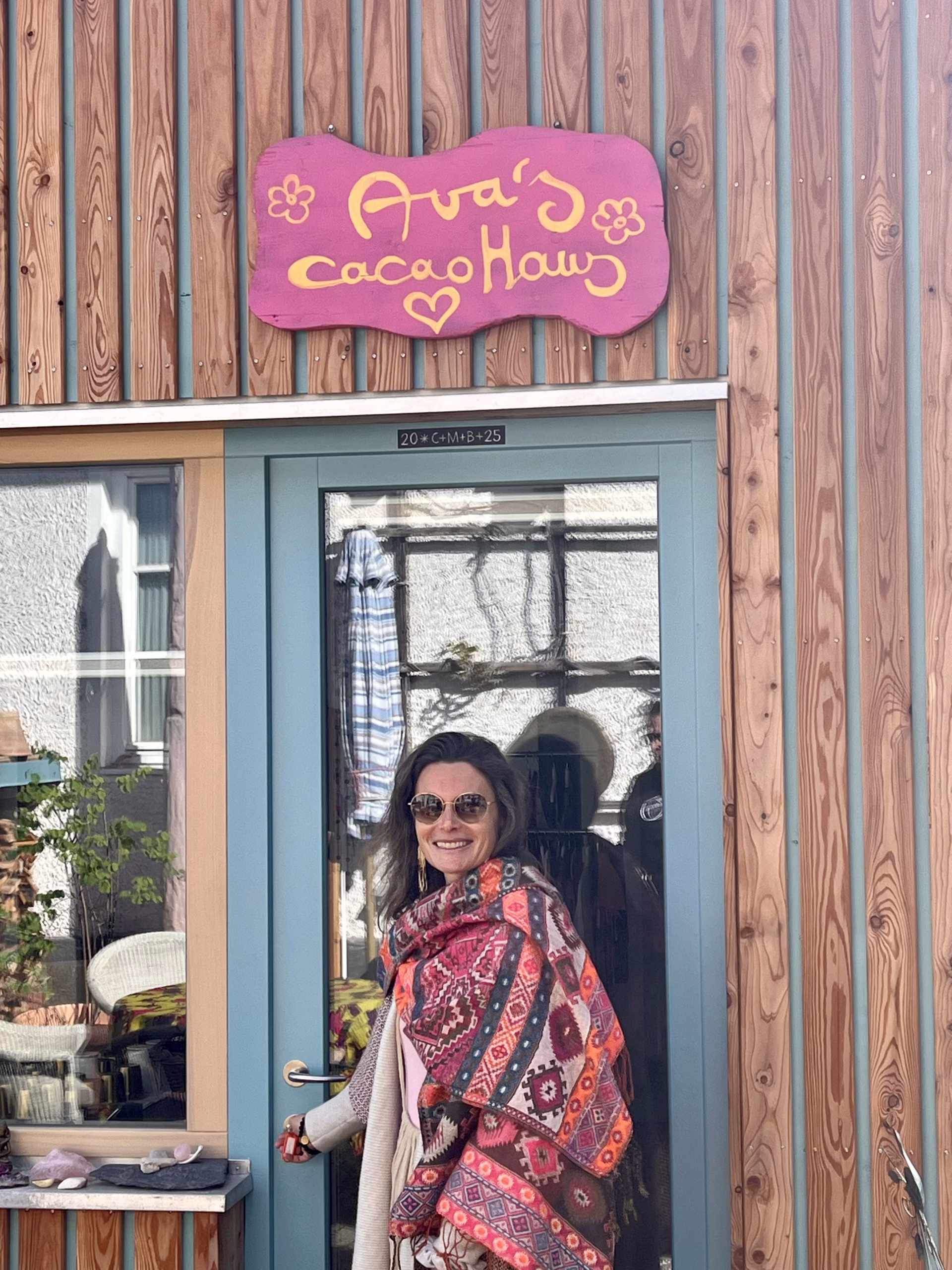Ava öffnet lächelnd die Tür zu ihrem cacaoHaus, einem einladenden Ort aus Holz mit handgemaltem Schild, Sinnbild für herzliche Begegnung und spirituelle Räume.